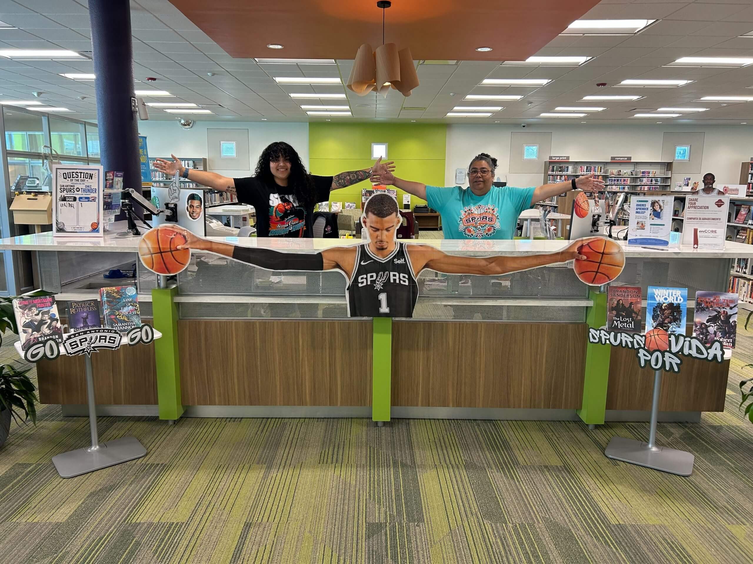Librarians Lila De Los Santos and Elvia Ramos stretch out their arms behind a cutout of Victor Wembanyama stretching out his. Both of them fit in his wingspan.