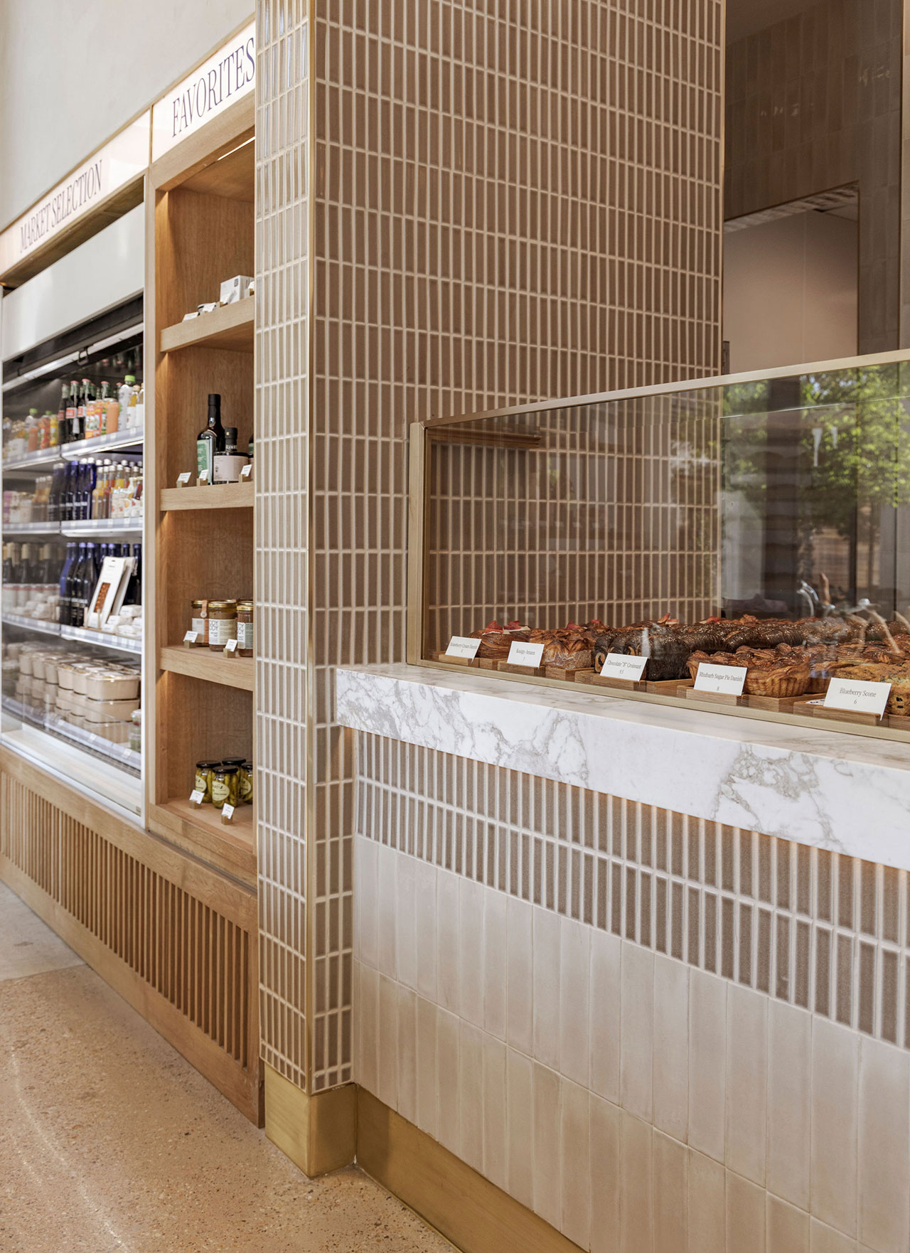 A bakery counter with pastries on display behind glass, shelves of packaged goods, and a tiled wall in a modern, minimalist interior.
