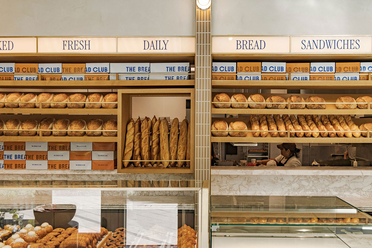 Bakery display with shelves of baguettes and round loaves, labeled “Fresh Daily Bread,” with a worker behind the counter and pastries in a glass case in the foreground.