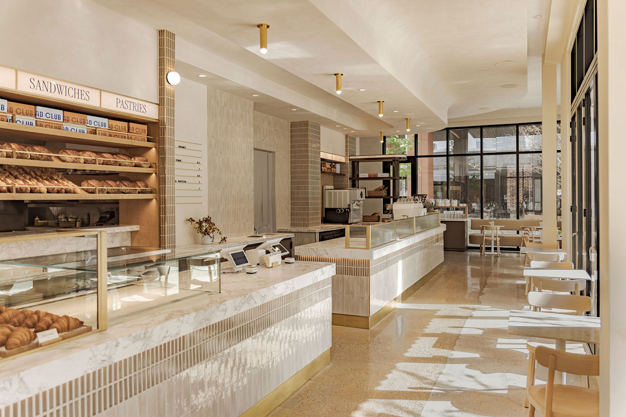 Modern bakery café interior with marble counters, wooden shelves displaying pastries, sandwich and pastry signs, and tables and chairs by large windows letting in natural light.