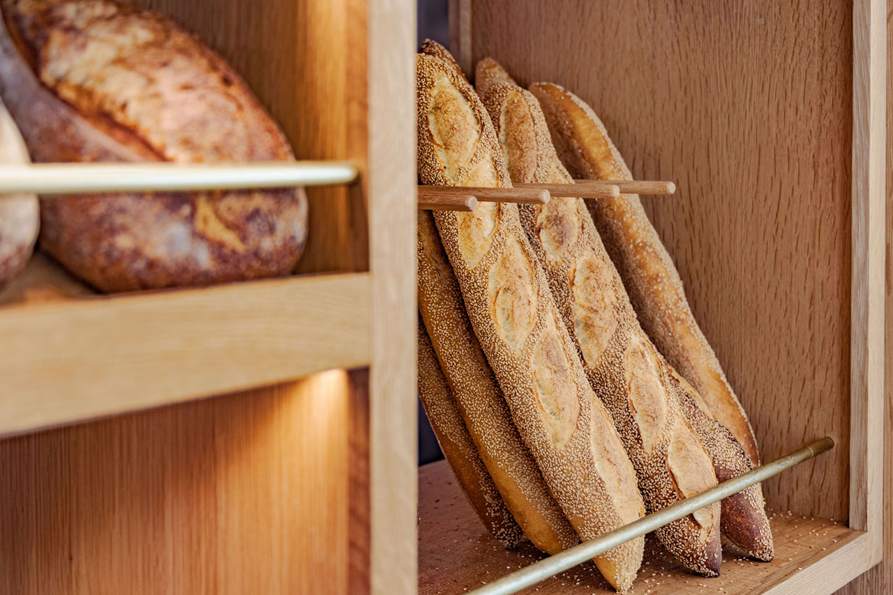 Several baguettes are displayed upright on a wooden shelf, separated by dowels, with other loaves of bread visible on the left.