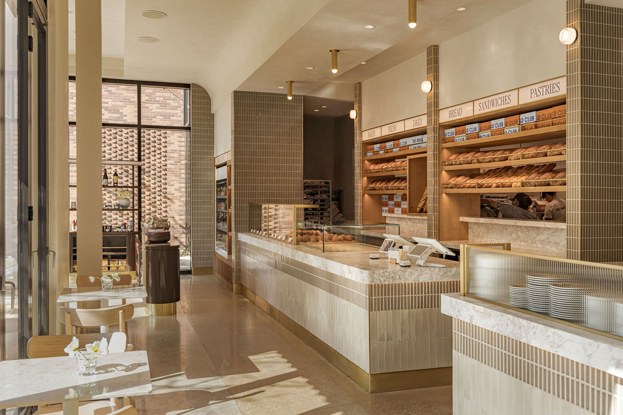 Modern bakery interior with tiled counters, display shelves filled with bread and pastries, and small tables with chairs by large windows letting in natural light.