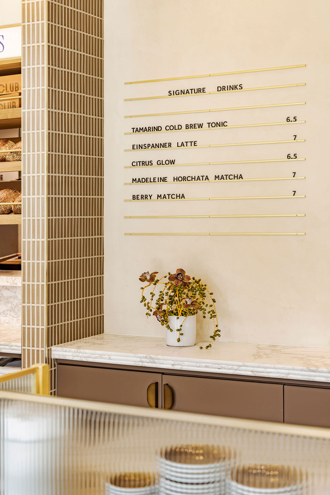 A countertop with a potted plant sits beneath a menu listing signature drinks and prices on a cream-colored wall in a modern café.