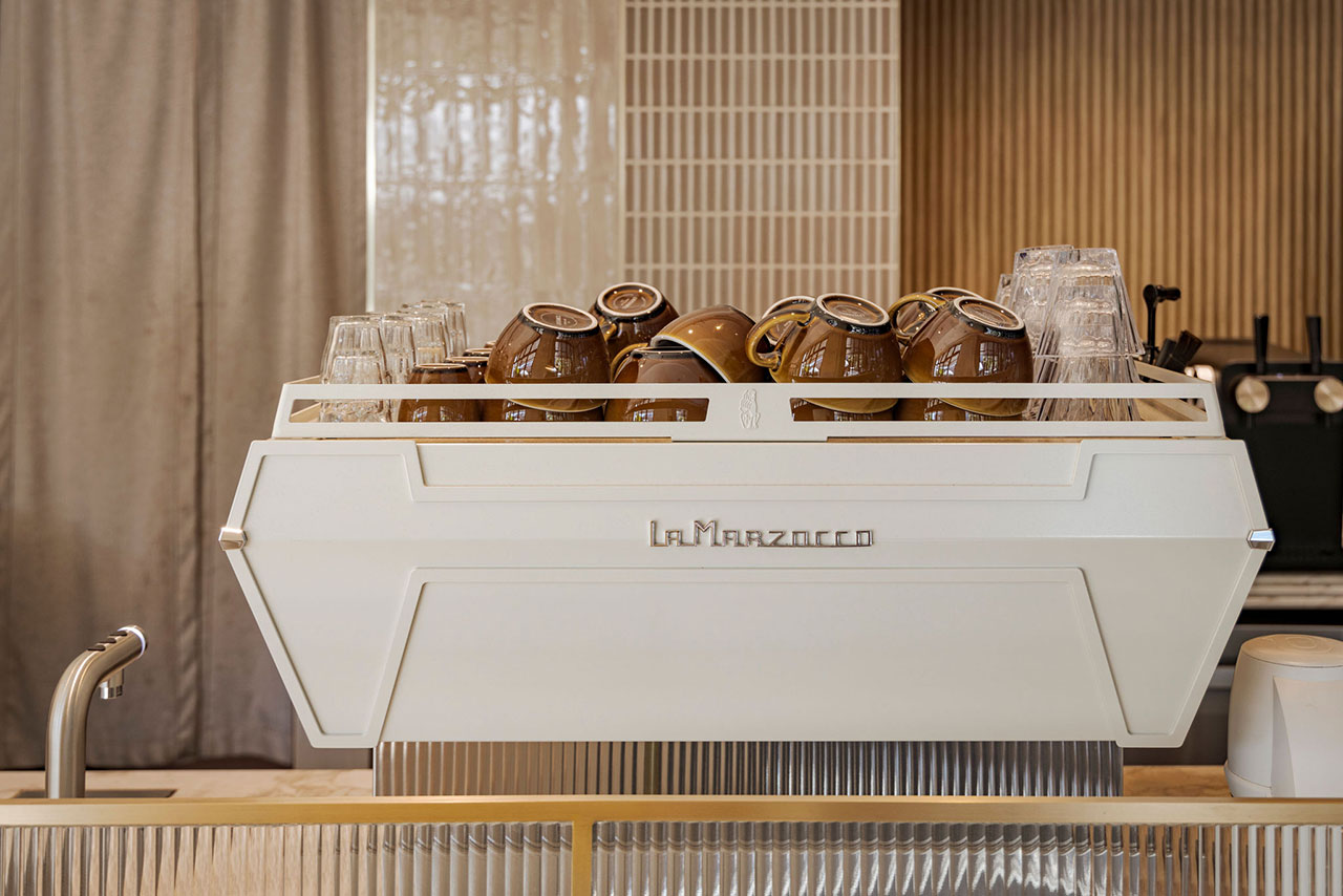 A white La Marzocco espresso machine with stacked brown cups and clear glasses on top, situated on a countertop in a modern cafe setting.