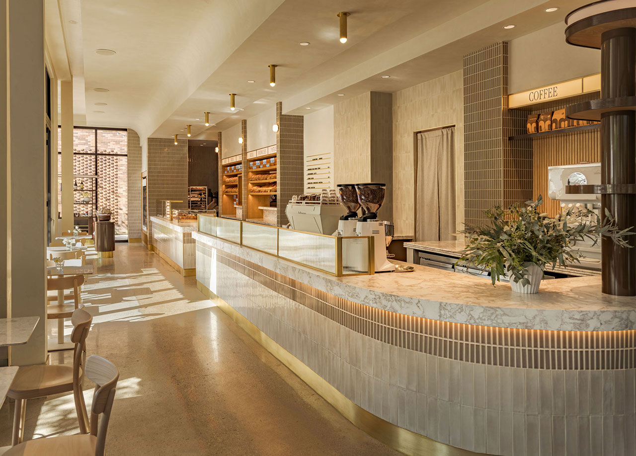 Modern coffee shop interior with a long marble counter, light wood furniture, coffee machines, and shelves with bread and pastries in the background.