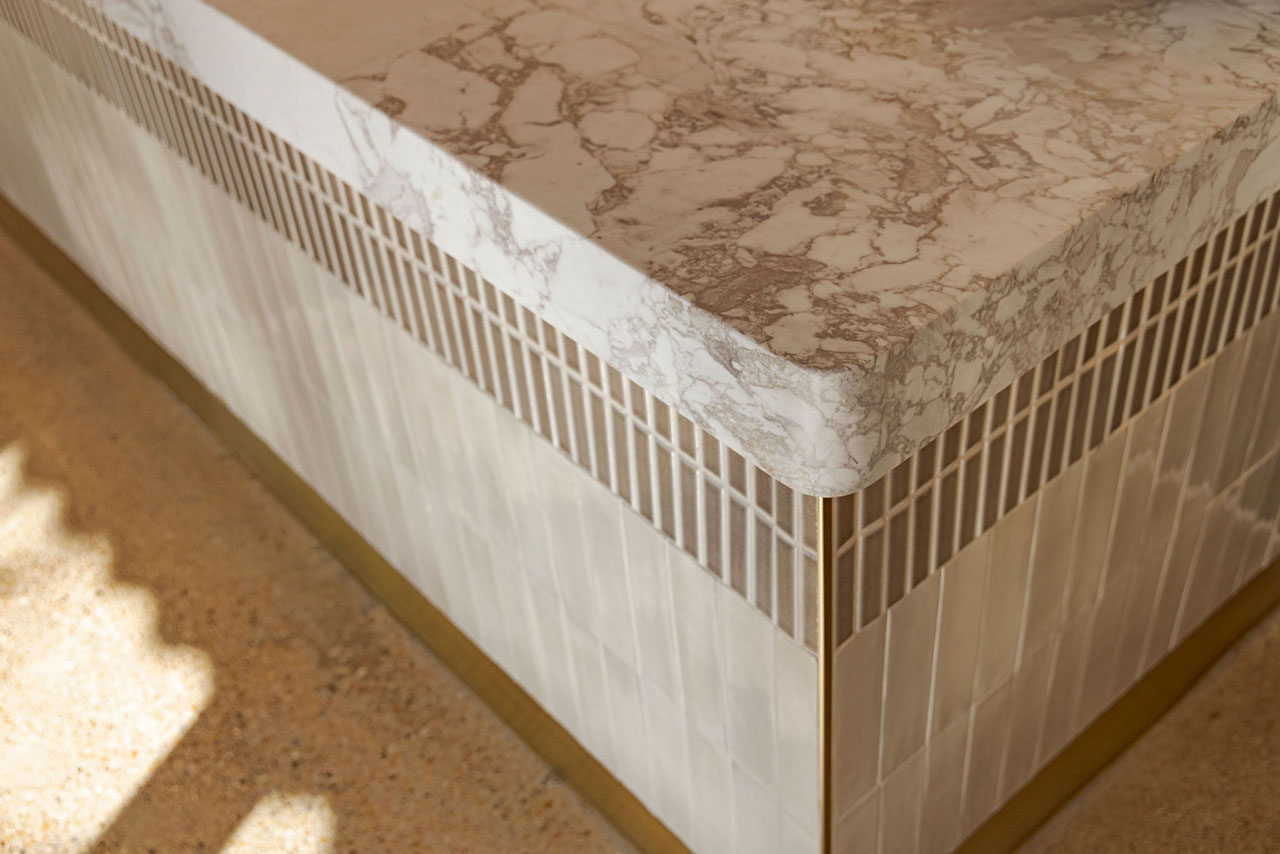 Marble countertop with rounded edge on a tiled base featuring vertical rectangular tiles and brass trim, set on a textured floor.