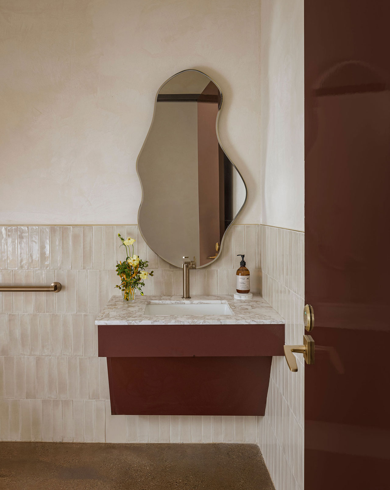 A bathroom with a marble-topped burgundy vanity, an irregularly shaped mirror, beige tiled walls, and a soap dispenser and small vase with flowers on the sink.