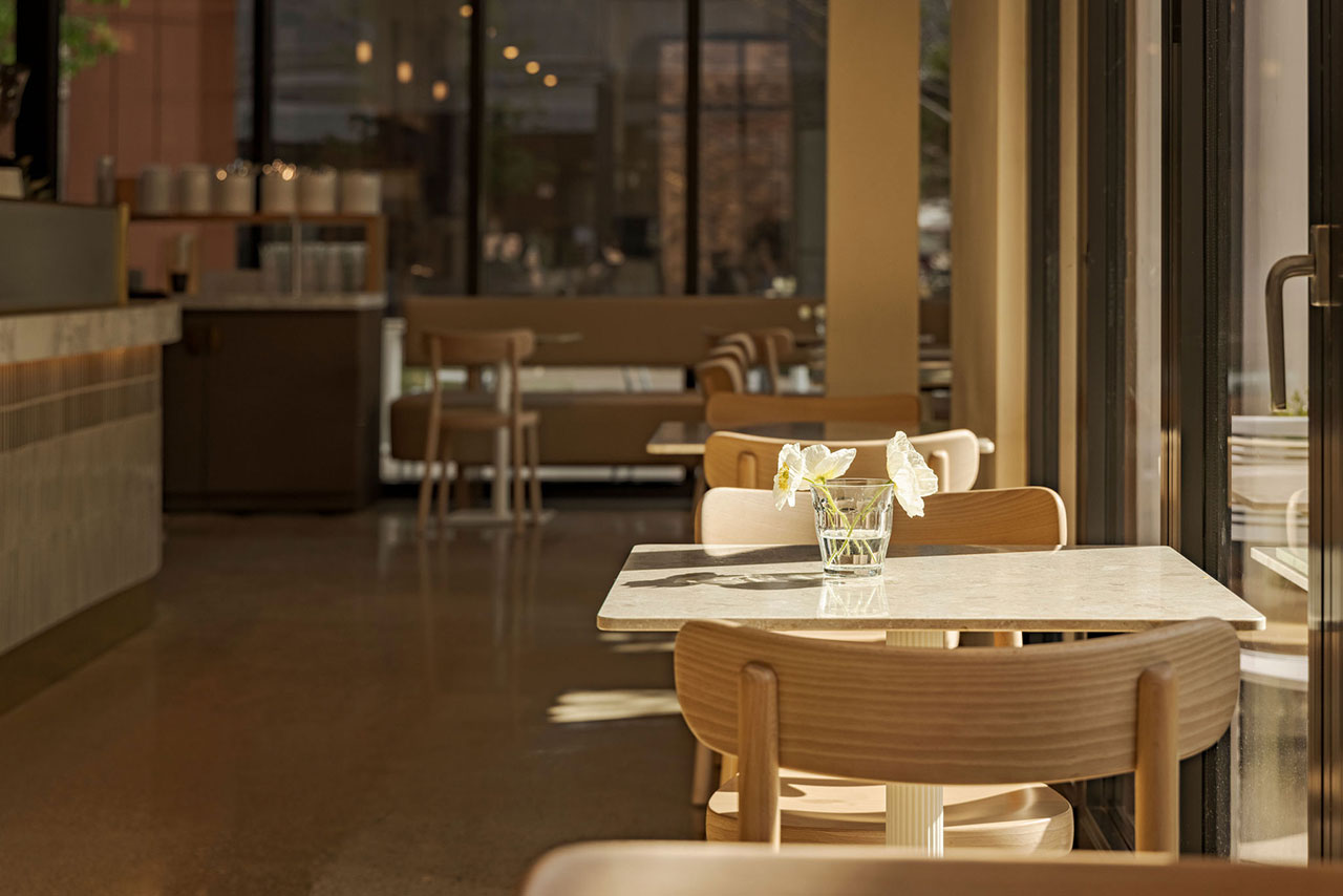 A sunlit cafe interior with wooden chairs and a marble table, featuring a glass vase with white flowers, near large floor-to-ceiling windows.