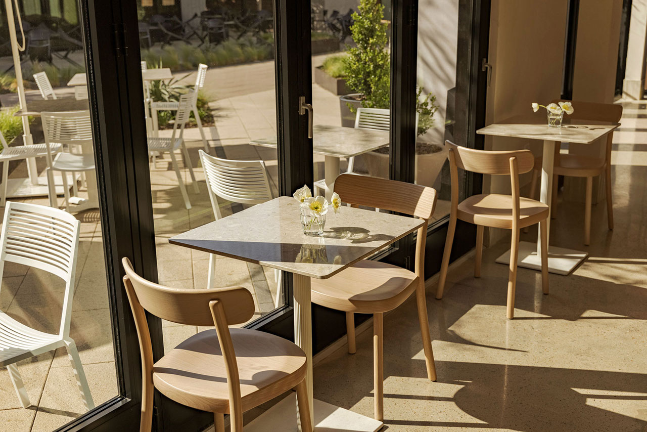 A sunlit café with wooden tables and chairs near large glass windows; each table has a small vase with white flowers. Outdoor seating is visible through the windows.