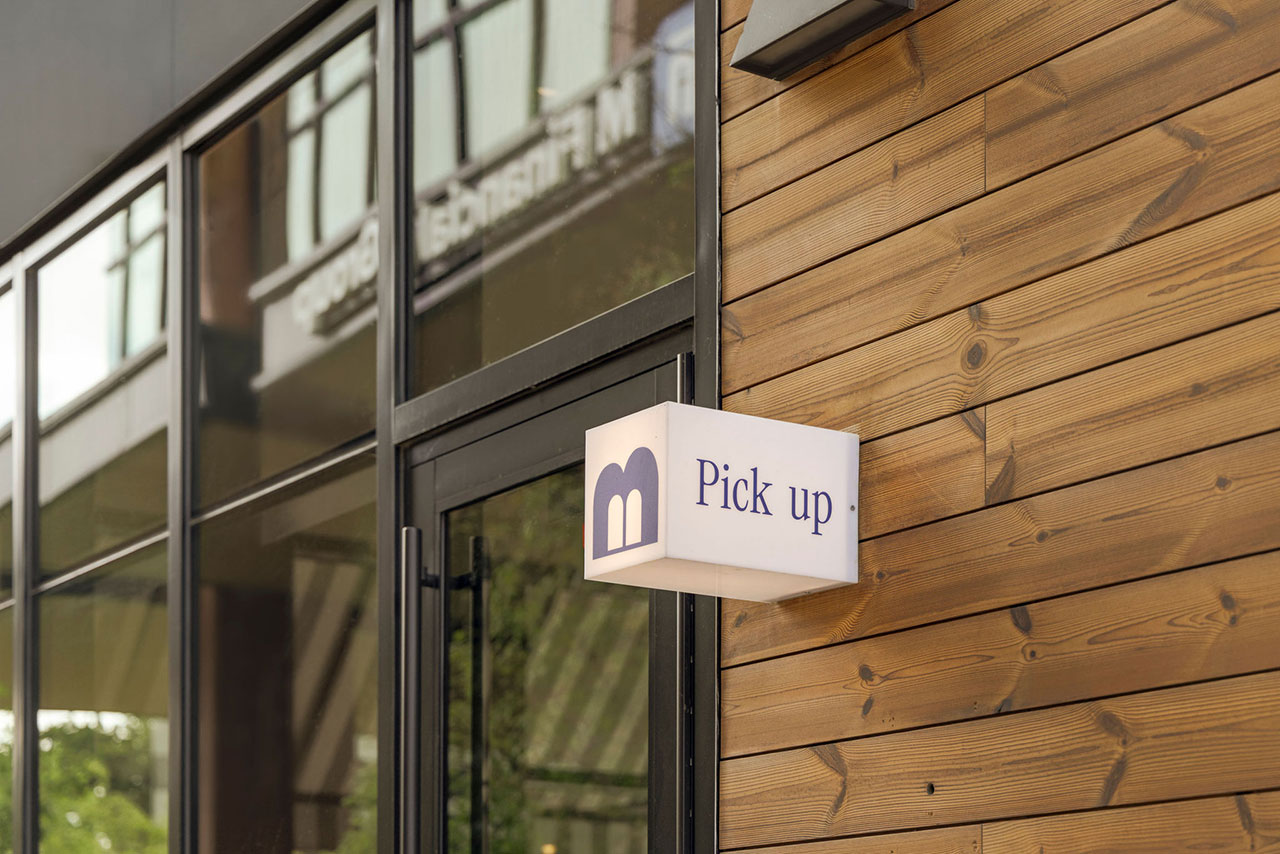 A sign reading "Pick up" with a logo is mounted on the exterior wooden wall of a modern building with large glass windows.