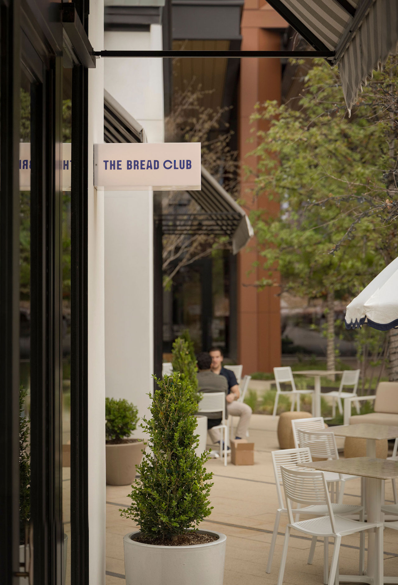 Outdoor seating area with white tables and chairs near potted plants, next to a building with a sign reading "The Bread Club." A person sits at a table in the background.