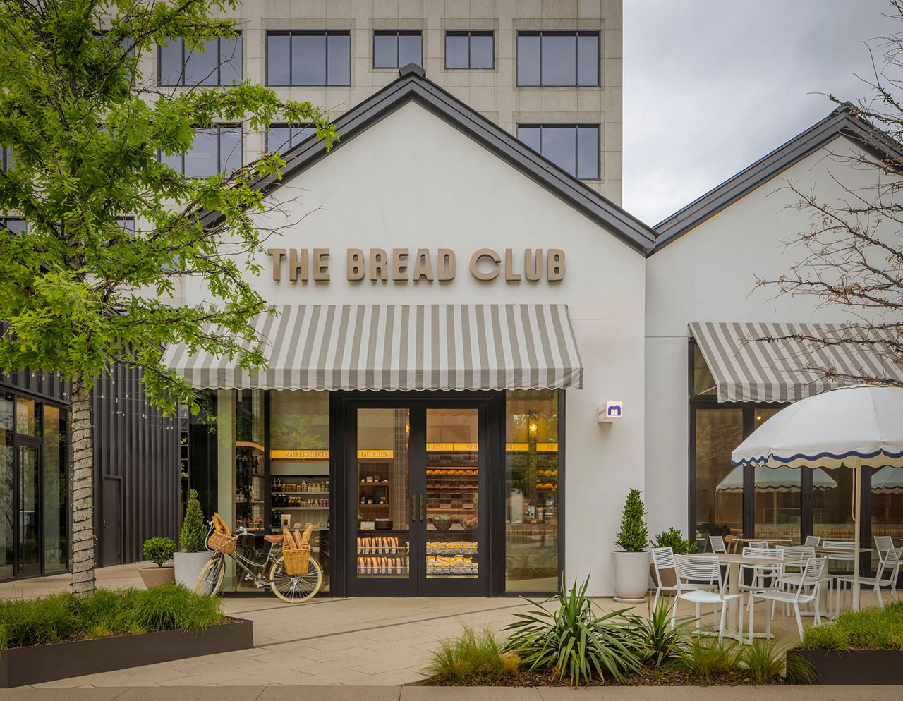A bakery called "The Bread Club" with striped awnings, outdoor seating, a bicycle with baskets, and large display windows under a cloudy sky.