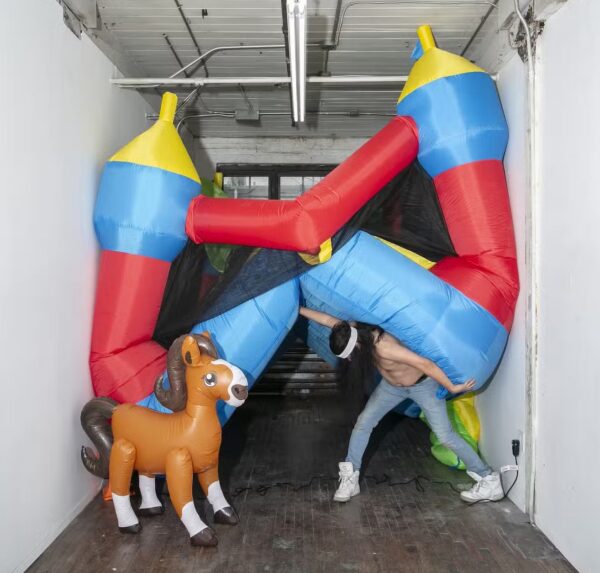 A shirtless man stands in a hallway holding a bounce house on his back resembling Atlas holding the world, with an inflatable horse staring at him.