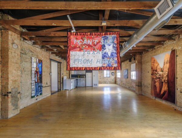 A gallery installation view of paintings on brick walls, with a central suspended textile artwork of an upside-down Texas flag with the words "I can't see Texas from here."