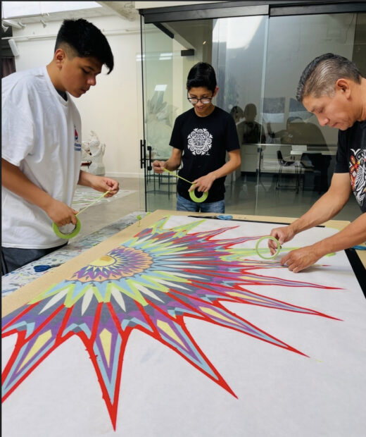 A photograph of students and an artist instructor using colorful tape to create a sunlike design on a large piece of paper.