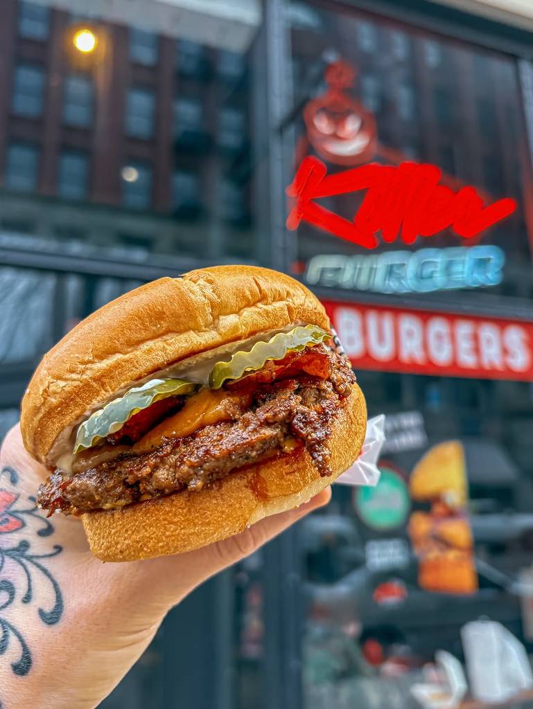 Image shows a person someone holding up a peanut butter bacon burger with the Killer Burger sign in the background.
