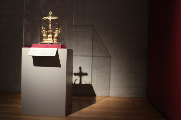 A dramatically lit installation image of a reliquary with a cross on top and flanked by two angels.