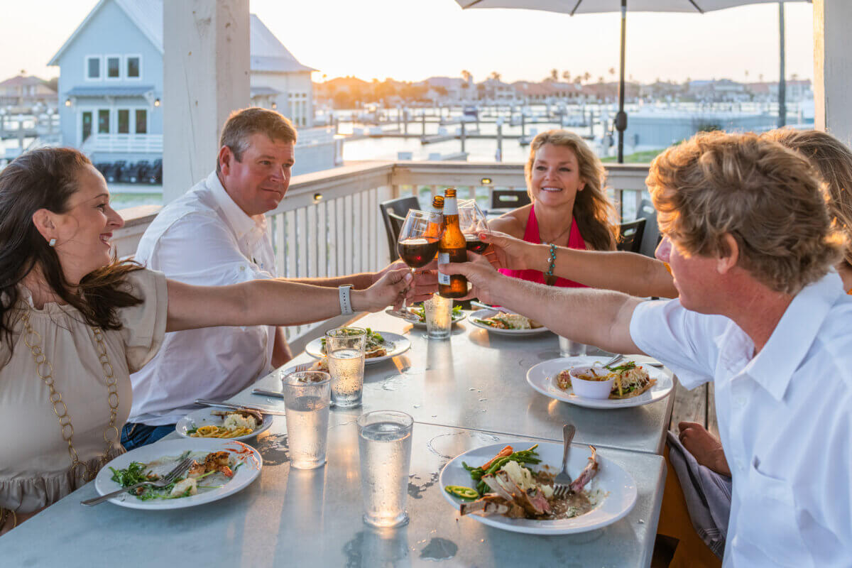 Five people seated at an outdoor table by the water clink their drinks together over plates of food at sunset—an unforgettable way to eat in Texas.