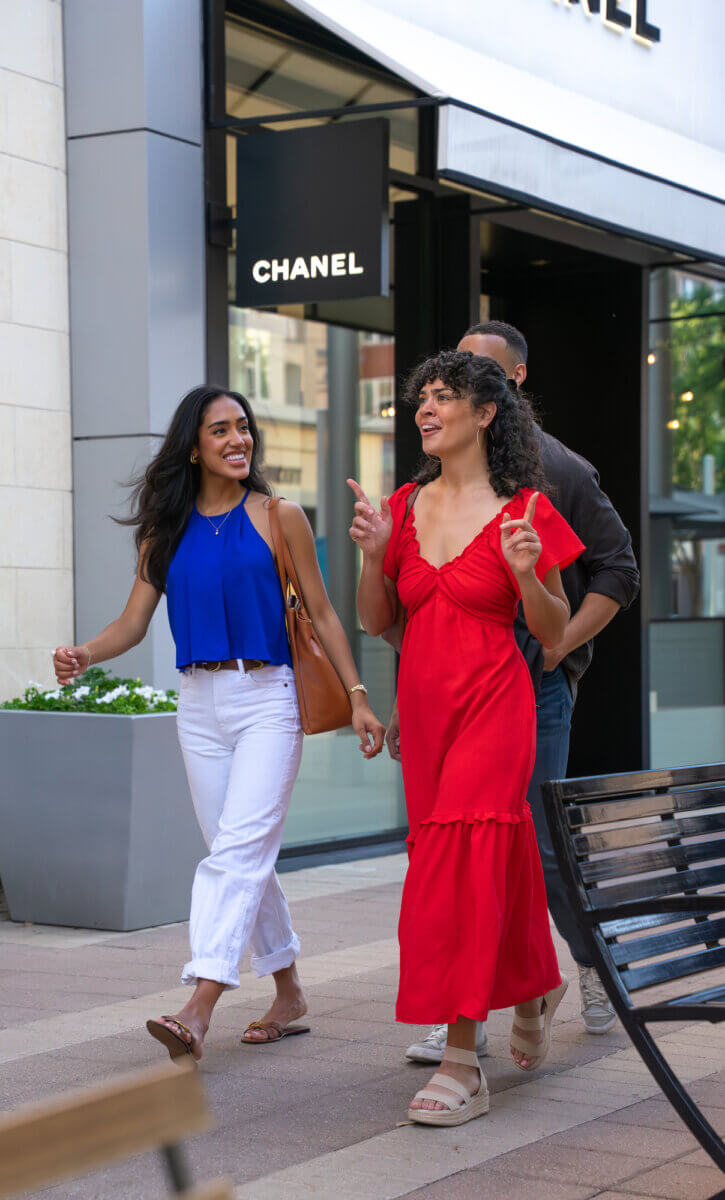 Three people walk and talk outside a Chanel store in Texas; one woman wears a red dress, another a blue top and white pants, with a man behind them.
