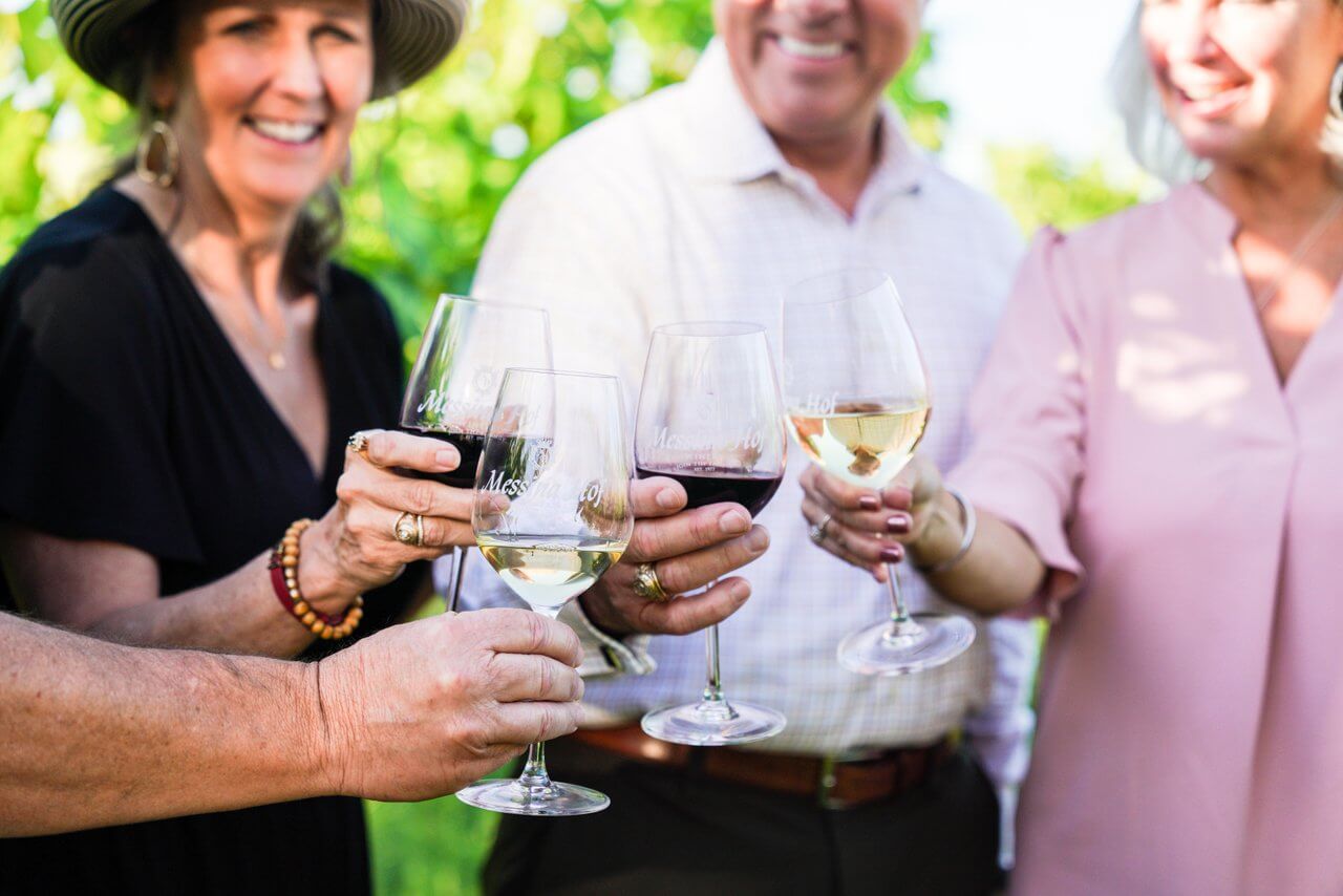Four adults smiling and clinking glasses of red and white wine outdoors, surrounded by greenery, celebrate the good life—just another reason to eat in Texas and savor unforgettable moments together.