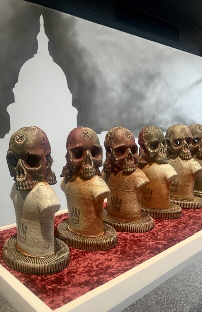 A close-up view of a table holding a row of skull-headed ceramic busts, in front of a wall painting of a silhouetted federal Capitol Building.