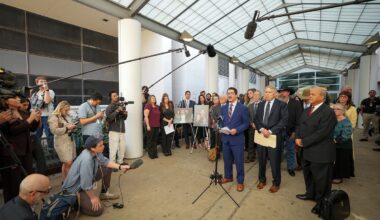 Travis County District Attorney José Garza, a Mexican man in a blue suit, speaks to reporters while a group of people stand behind him with portraits on printed boards.