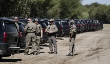 A group of officers stand on a dirt road behind a line of SUVs.