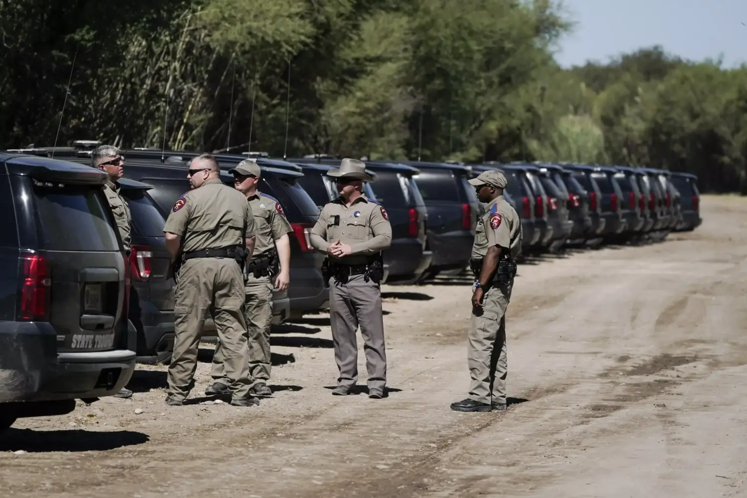 A group of officers stand on a dirt road behind a line of SUVs.
