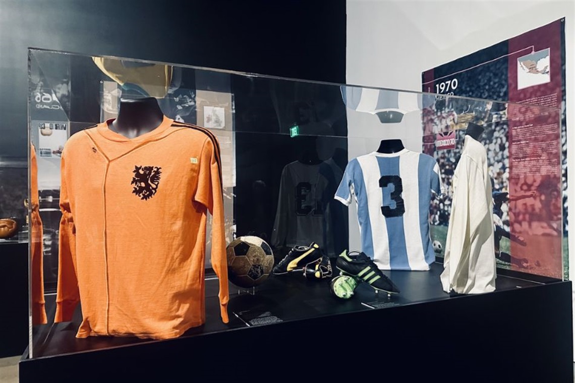 A display case with vintage soccer memorabilia, including an orange jersey with a lion crest, an Argentina jersey with the number 3, an old soccer ball, gloves, and boots.