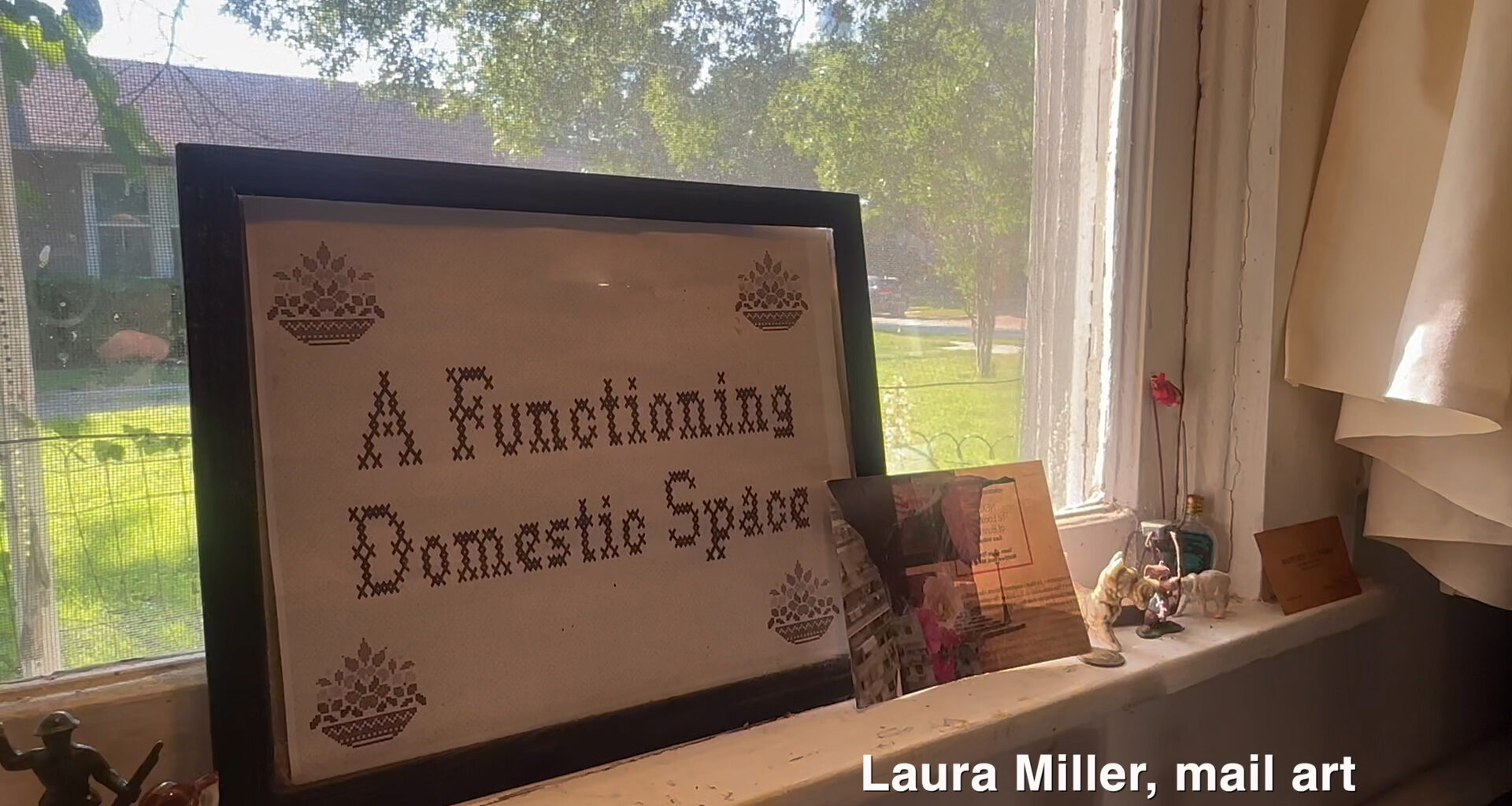 A framed cross-stitch piece reading “A Functioning Domestic Space” sitting on a windowsill.