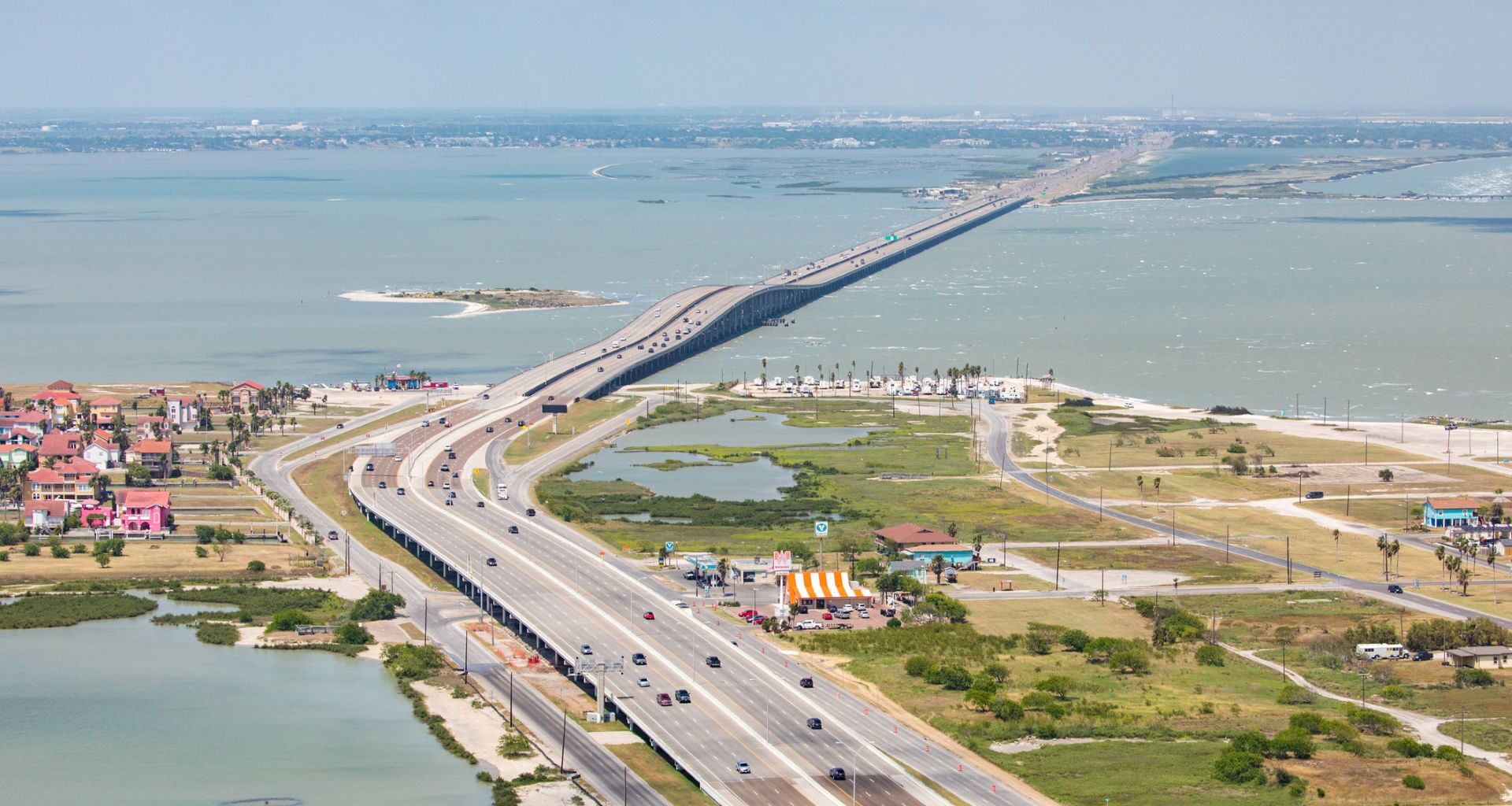 Aerial view of a highway bridge connecting two coastal areas, with water and green landscapes nearby.