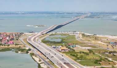 Aerial view of a highway bridge connecting two coastal areas, with water and green landscapes nearby.
