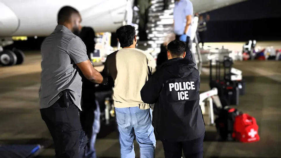 ICE personnel escort a male detainee in hand restraints up stairs to board a deportation plane