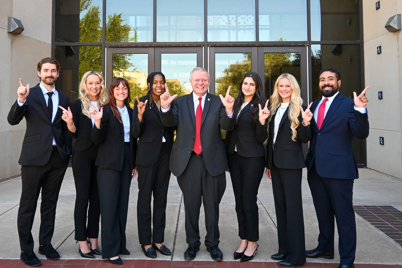 Nine people dressed in professional black suits stand in a row outside the Texas Tech School of Law building, all holding up the Guns Up hand sign. A man in a black suit with a red tie stands at the center of the group.