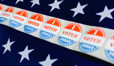 A photographic illustration depicting a strip of I voted stickers over two rows of whites stars over a blue background.