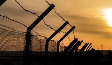 A photograph of a fence topped with barbwire in front of an orange sunset.
