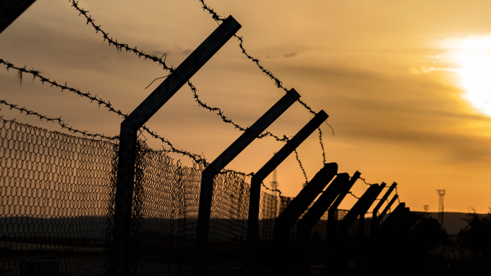 A photograph of a fence topped with barbwire in front of an orange sunset.