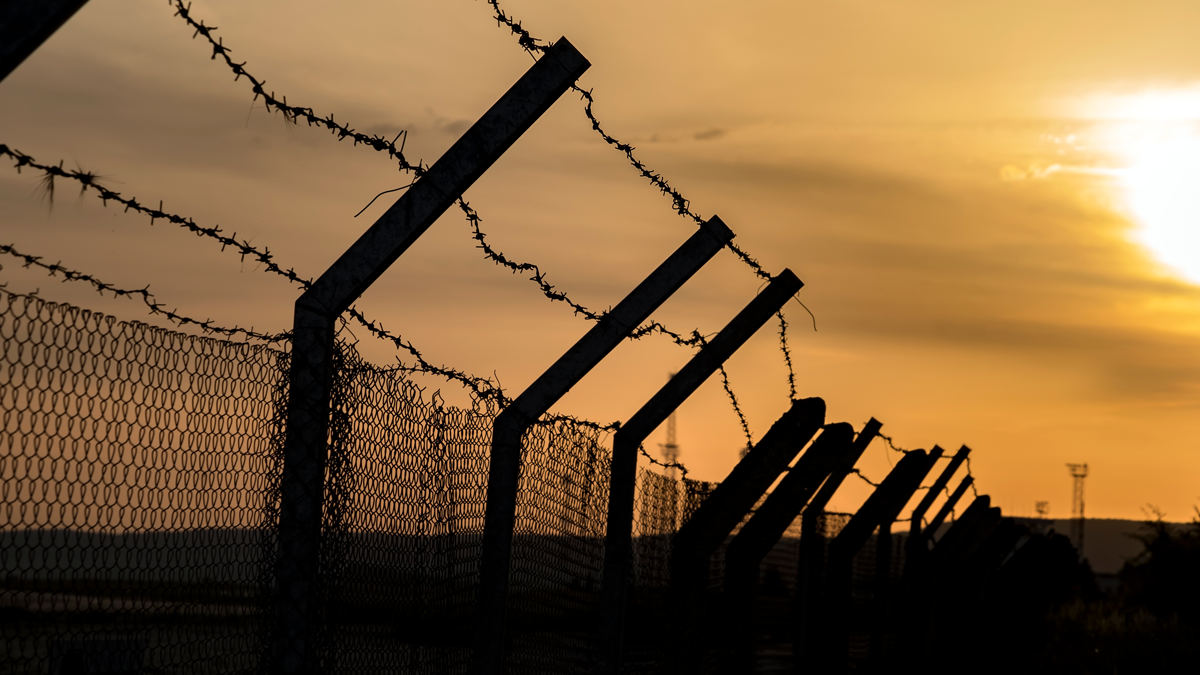 A photograph of a fence topped with barbwire in front of an orange sunset.
