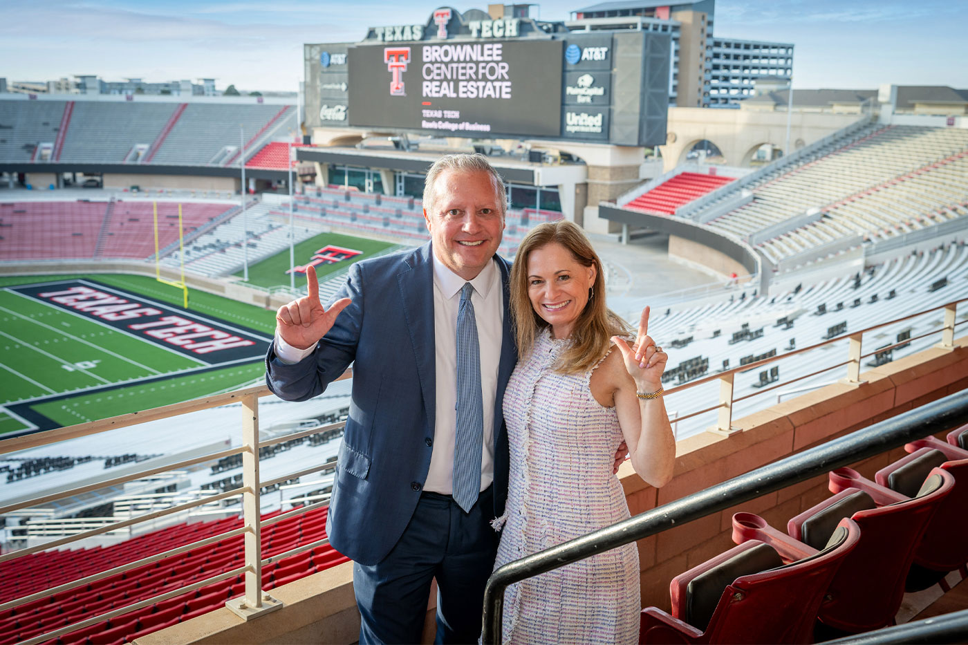 John and Rachel Brownlee stand together in the stands of Jones AT&T Stadium, both holding up the Guns Up hand sign and smiling. The stadium scoreboard behind them displays "Brownlee Center for Real Estate, Texas Tech Rawls College of Business.