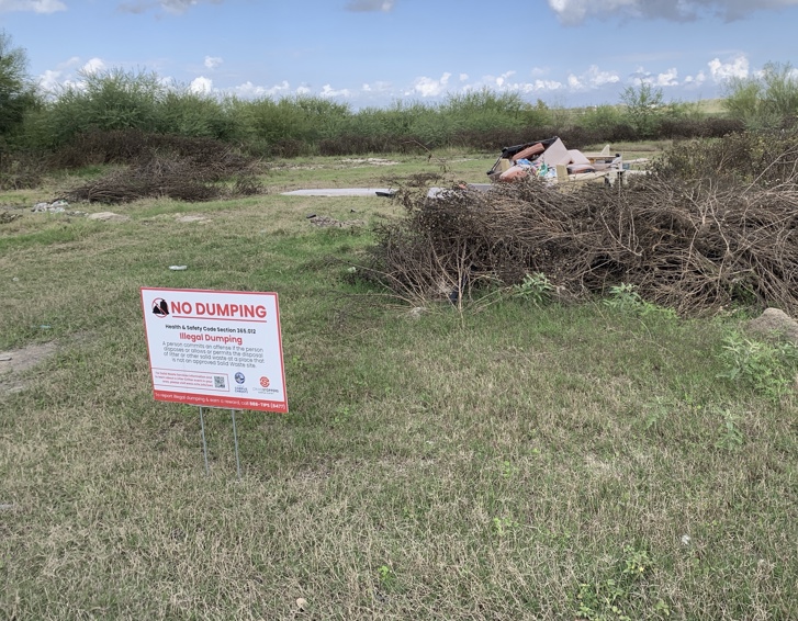 A new "No Dumping" sign is visible in a grassy area with debris including discarded furniture in the background.