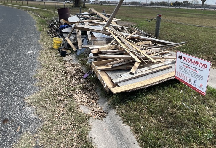 A pile of construction material and debris is dumped along the roadside. A new "No Dumping" sign is placed by the pile in the grass.