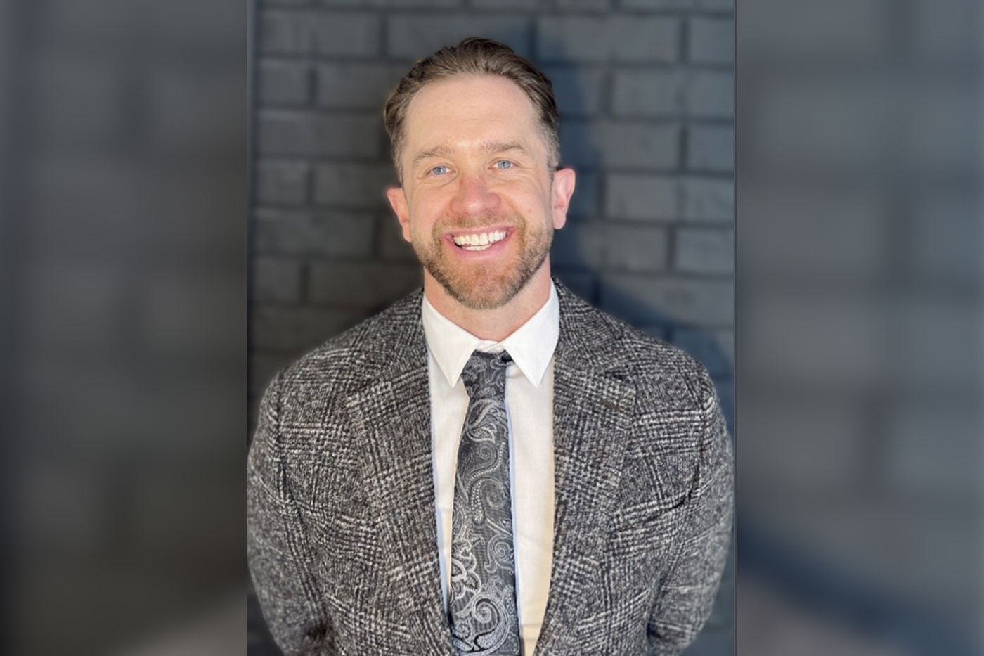 A man wearing a patterned jacket and tie, Seth Porter, smiles for a portrait against a dark brick background.