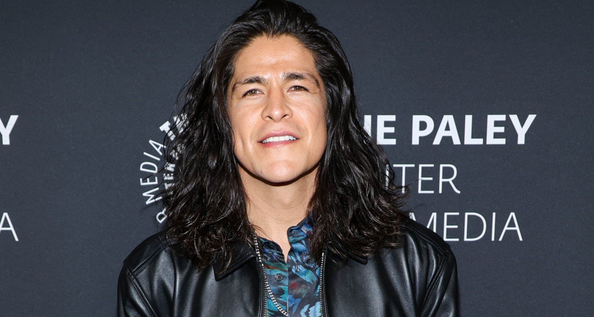 Cristo Fernandez with long dark hair wearing a leather jacket stands in front of a backdrop with text.