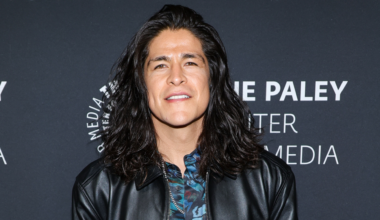 Cristo Fernandez with long dark hair wearing a leather jacket stands in front of a backdrop with text.