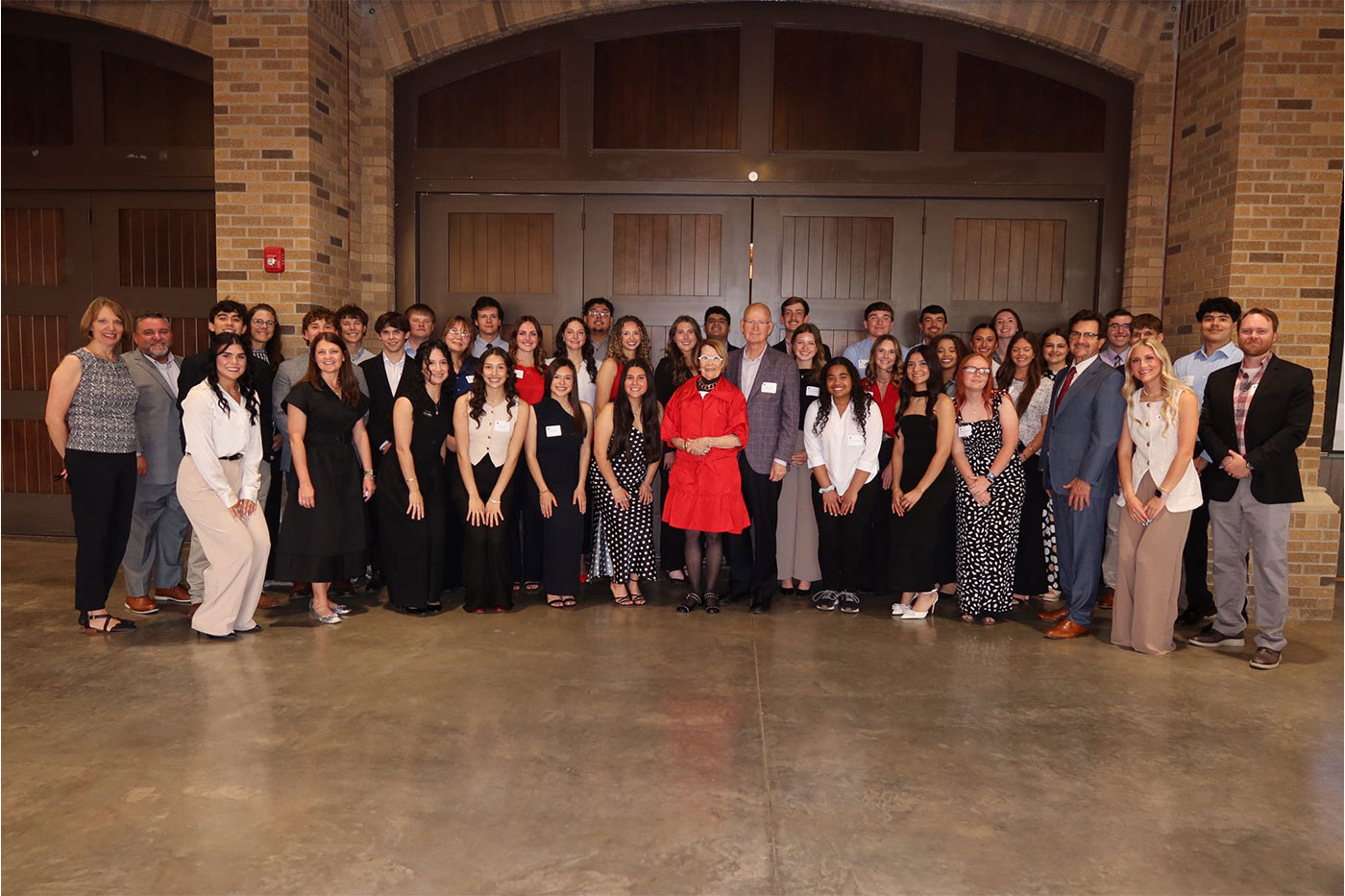 Approximately 40 people pose for a group photo inside a brick-walled event venue. Carl and Marilynn Thoma stand at the center, flanked by Texas Tech President Lawrence Schovanec and surrounded by Thoma Scholars dressed in formal attire.