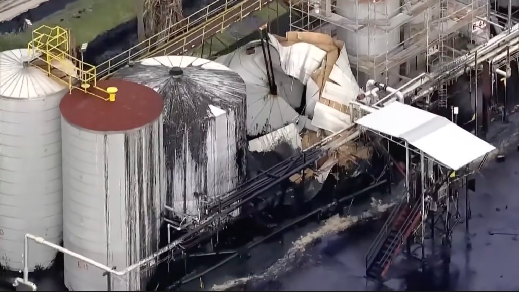 An industrial facility shows a ruptured asphalt tank, leaking black asphalt onto the ground, and two intact tanks with dark asphalt streaming down their sides.