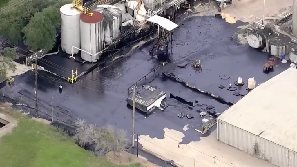 Aerial view of an industrial facility with dark liquid covering the ground, damaged tanks, and debris.