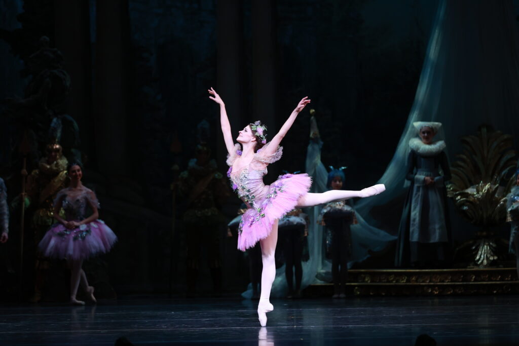Onstage, Jacquelyn Long does a piqué arabesque on pointe with her arms extended in a high "V." She wears a lilac-colored classical tutu with floral decoration and a tiara. 