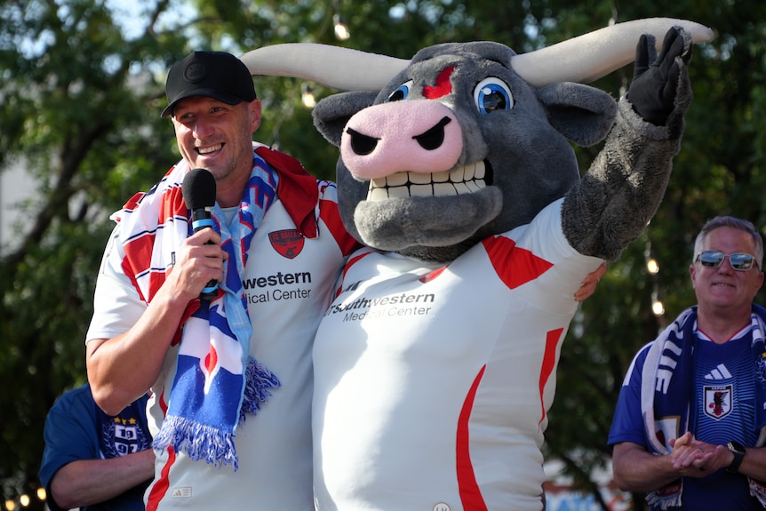 A white man wears a black baseball cap and hugs a mascot.