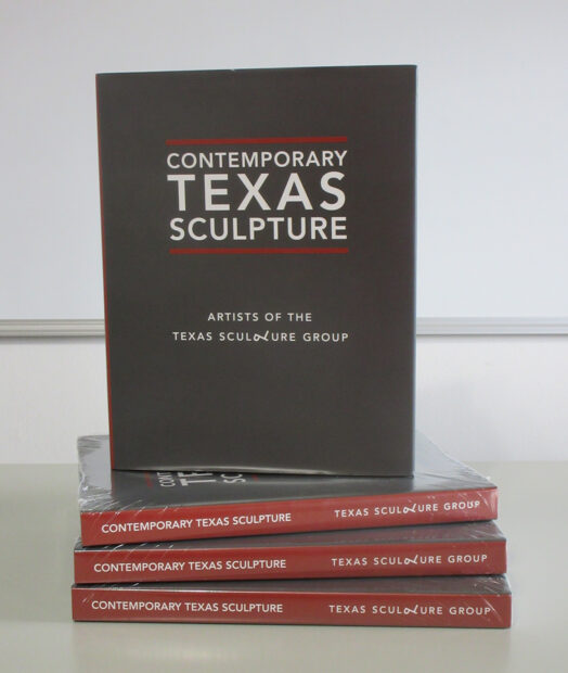 A photograph of a copy of the book “Contemporary Texas Sculpture: Artists of the Texas Sculpture Group” standing on top of three horizontally placed copies of the book. 